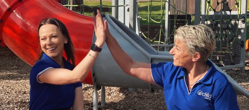 Two Child Australia staff members high fiving and smiling in front of an outdoor playground with a red slide, the sun is shining