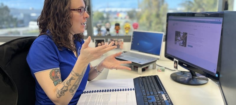 Staff member in a sunny office, with her arms raised in expression as she talks to someone on her computer