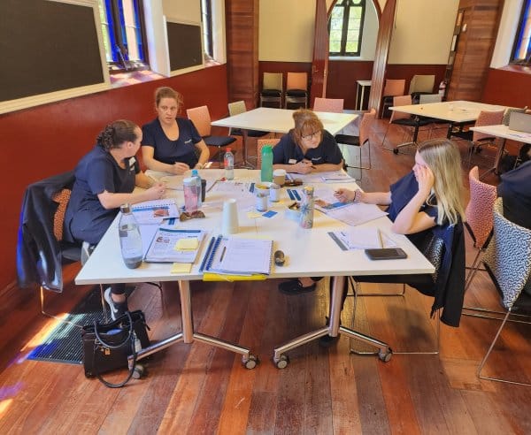 A group of women sitting at a desk covered with paper and books, writing and talking