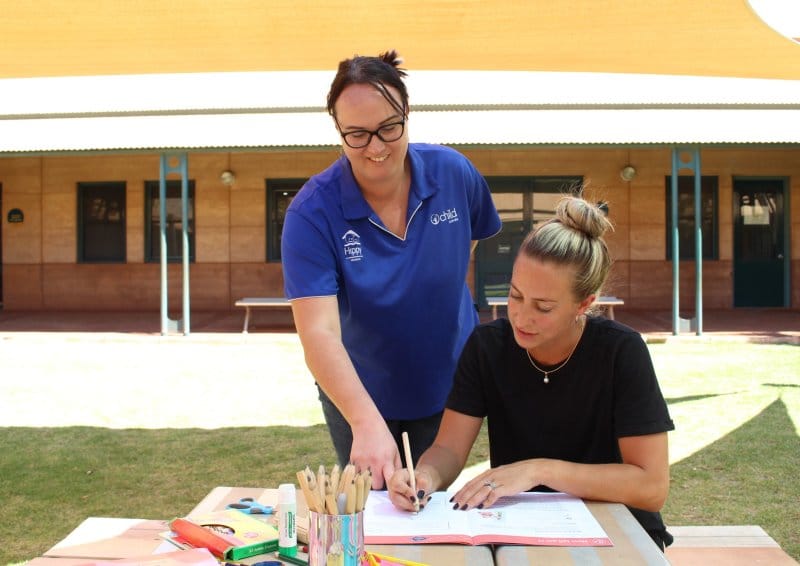 A woman writing on a notepad outside whilst another woman stands over her shoulder and points at a book