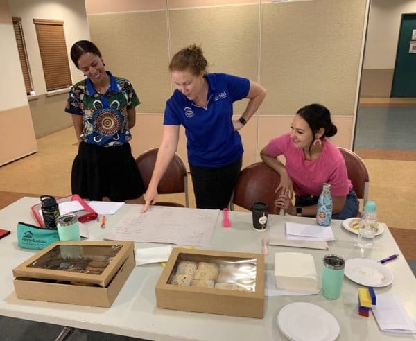 3 women at a desk with food and papers planning and talking.